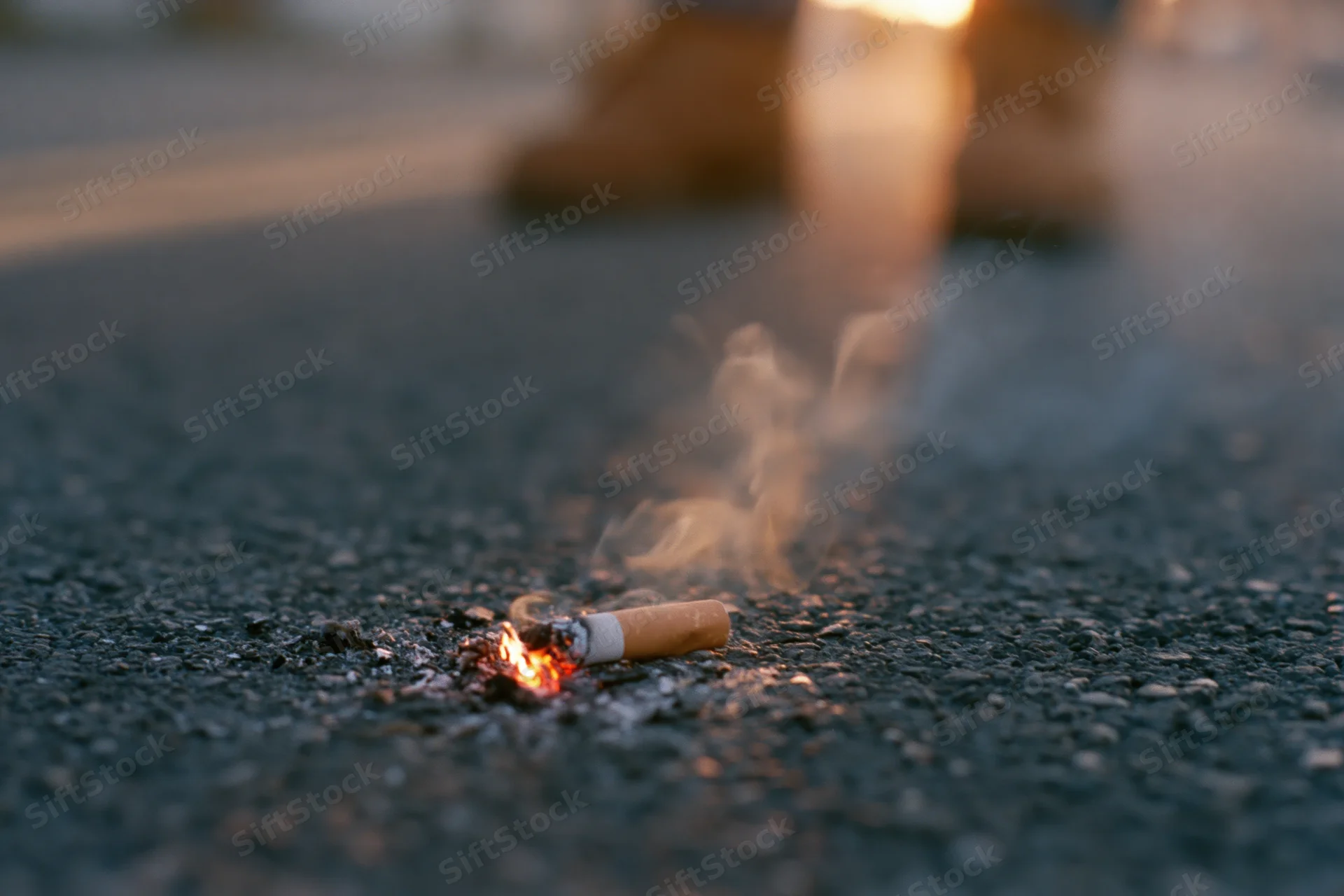 Lit cigarette butt stubbed out on dark asphalt pavement with smoke rising, feet blurred in background at dusk