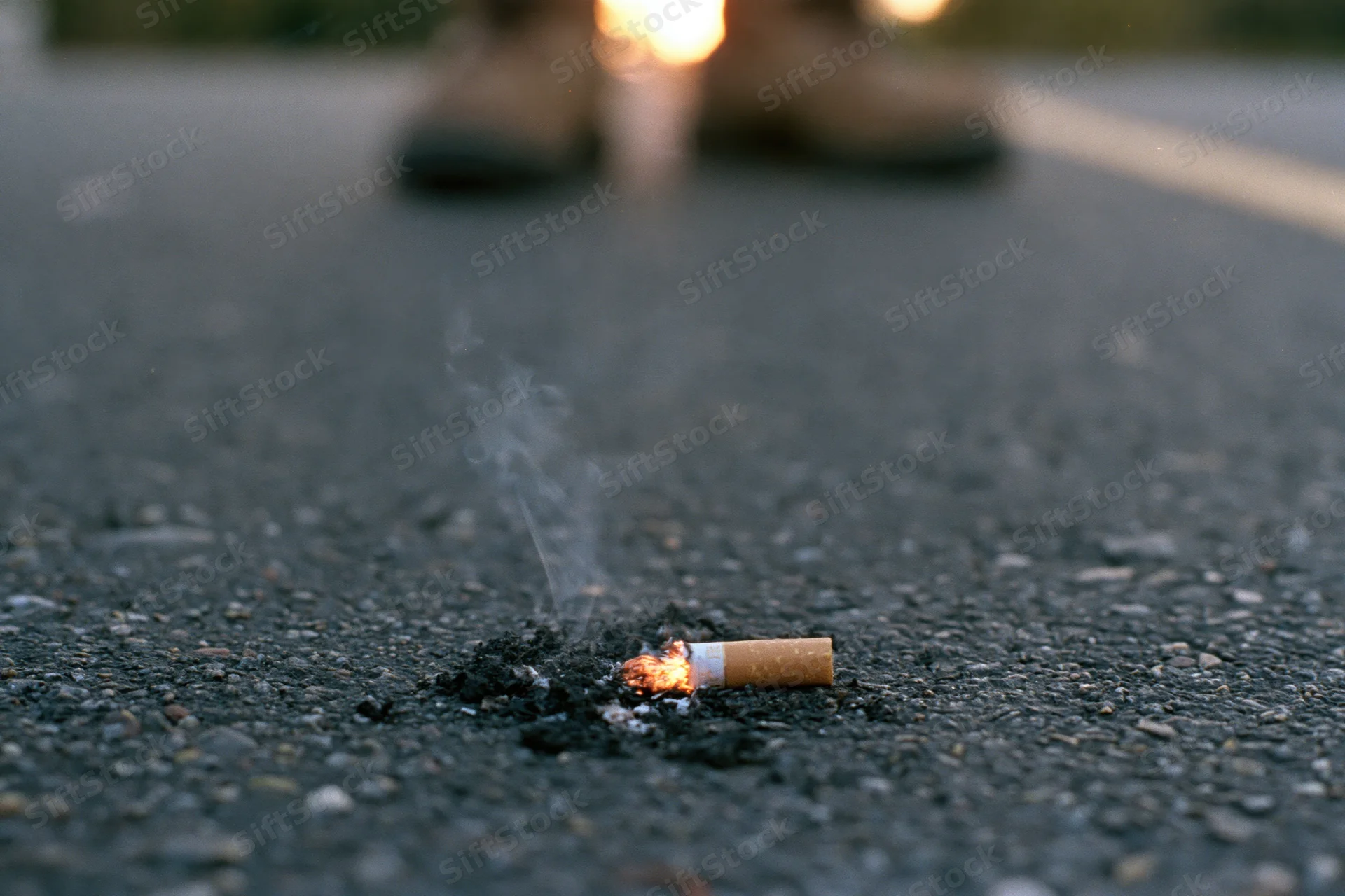 Lit cigarette butt smoldering on dark asphalt pavement with smoke rising and blurred shoes in background