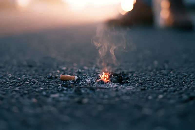 Lit cigarette butt smoldering on dark asphalt pavement with smoke rising, low-angle closeup view