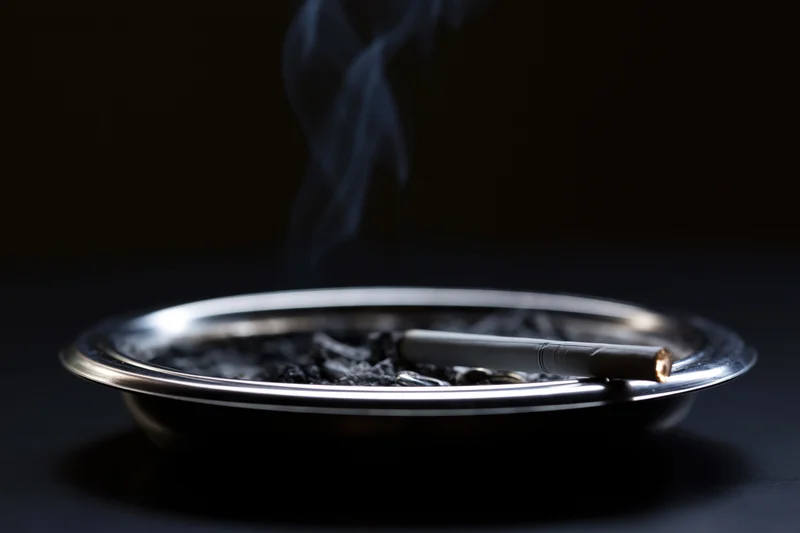 Lit cigarette resting in metal ashtray with rising smoke against dark background