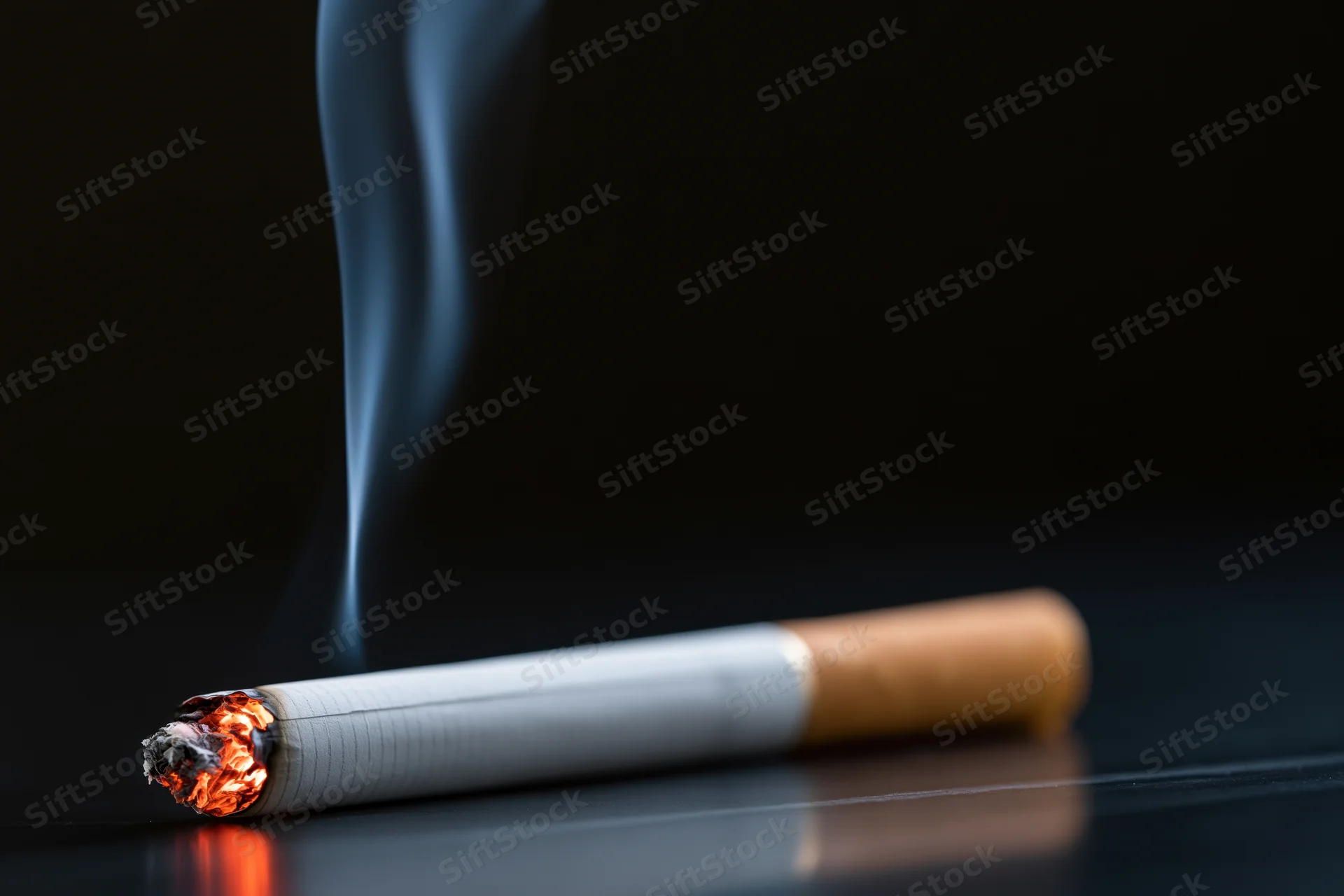 Lit cigarette burning on dark reflective surface with rising smoke