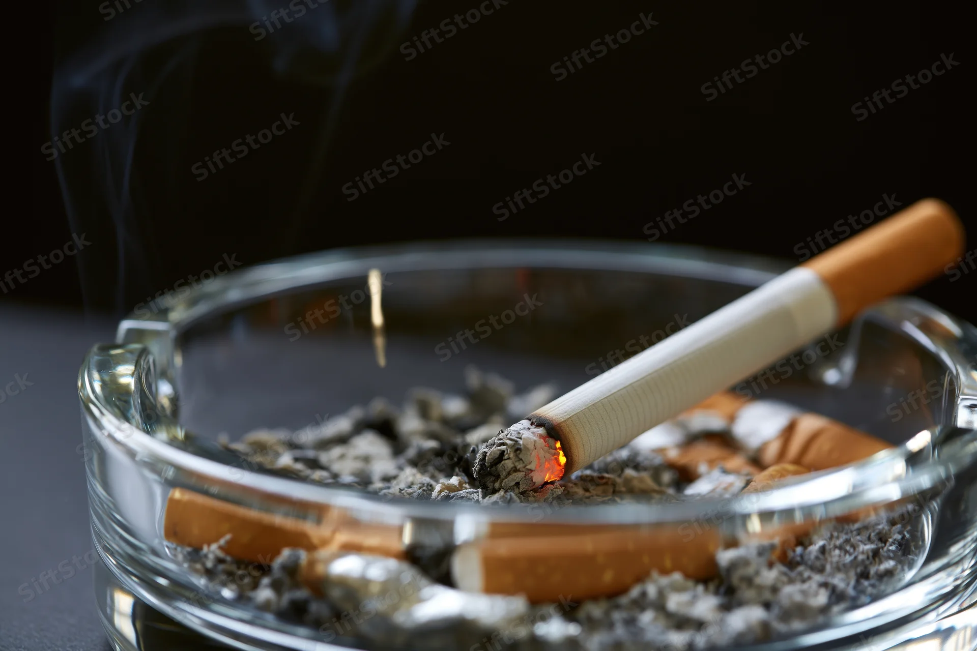 Lit cigarette burning in a clear glass ashtray filled with ash and cigarette butts