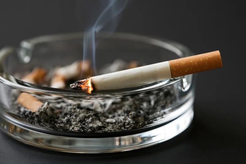 Lit cigarette burning in a clear glass ashtray filled with ash and cigarette butts on a dark surface
