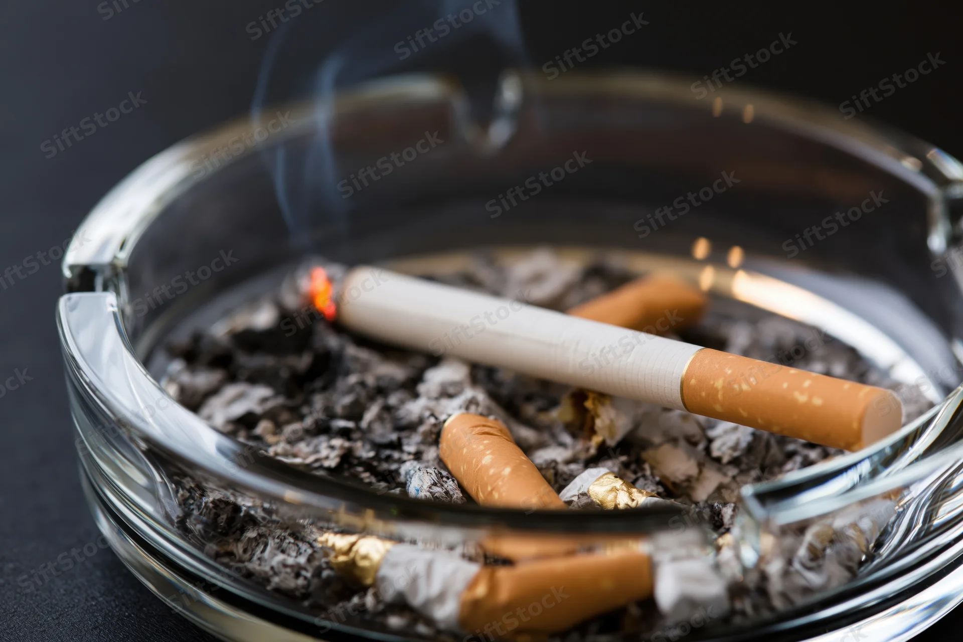 Lit cigarette burning in a clear glass ashtray filled with ash and cigarette butts on a dark surface