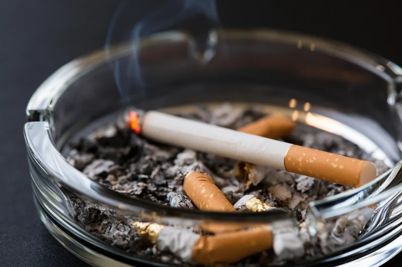 Lit cigarette burning in a clear glass ashtray filled with ash and cigarette butts on a dark surface