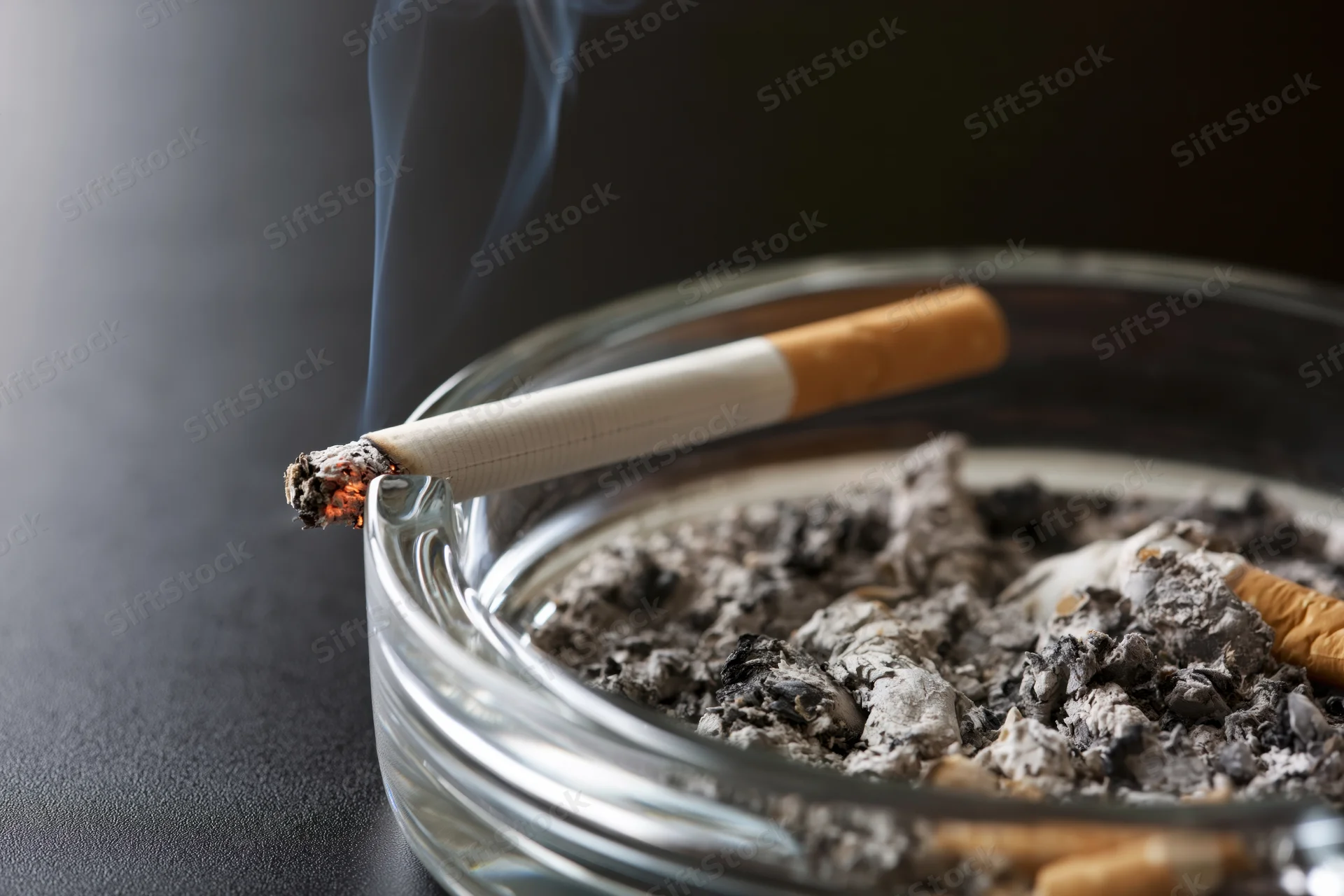 Lit cigarette burning in a glass ashtray filled with ash and cigarette butts on a dark surface