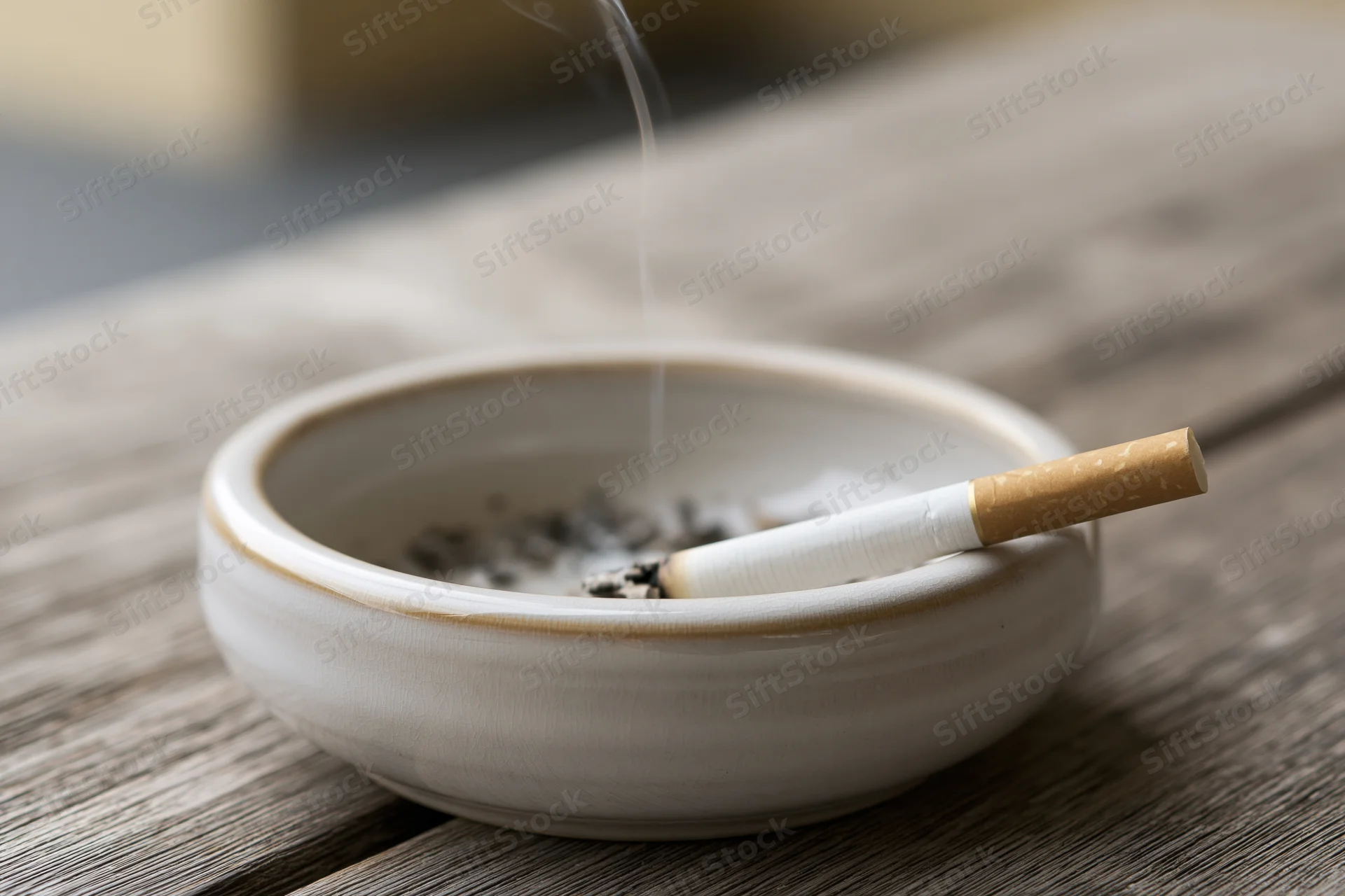 Lit cigarette resting in white ceramic ashtray on wooden table with smoke rising