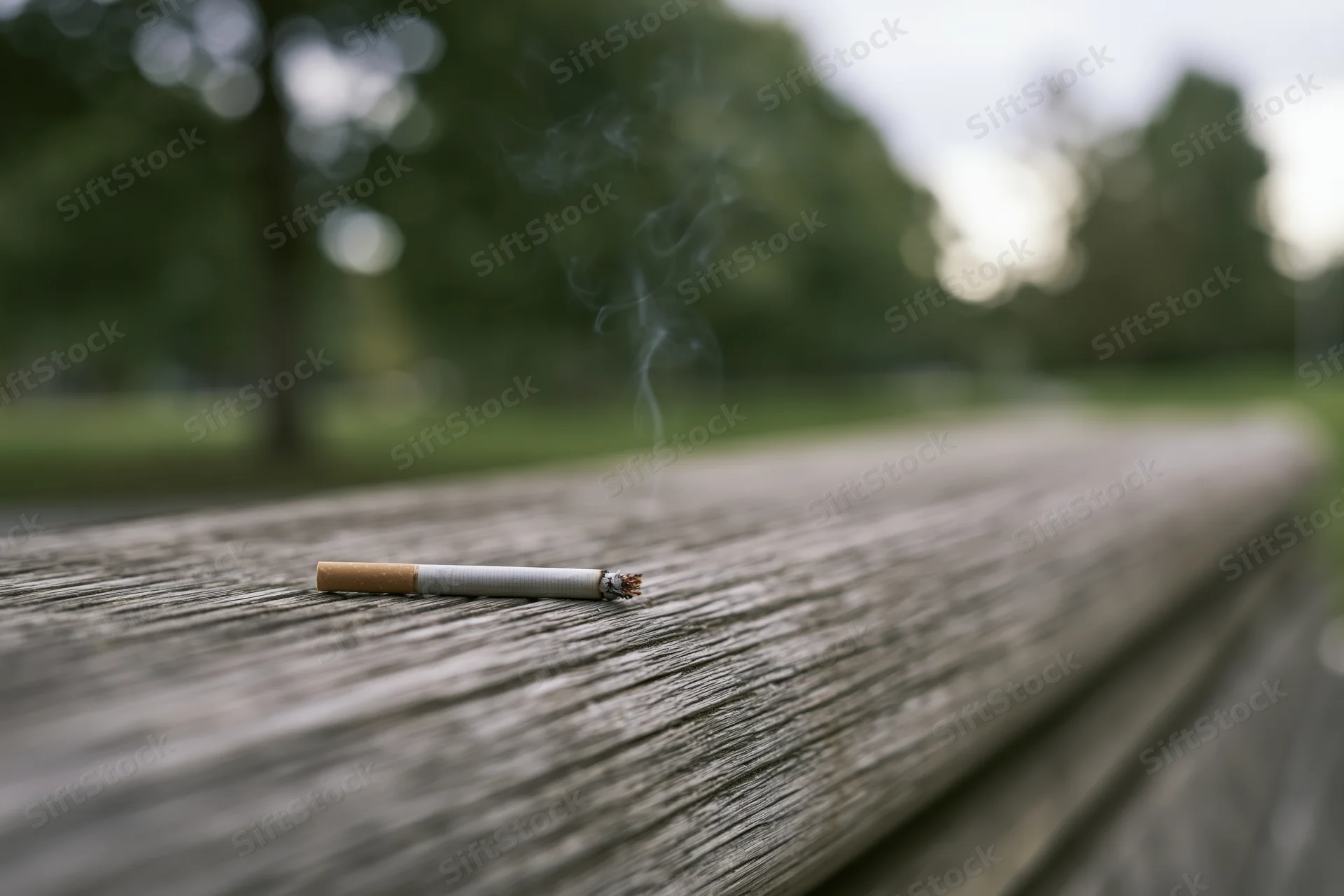Lit cigarette with smoke resting on weathered wooden park bench surface