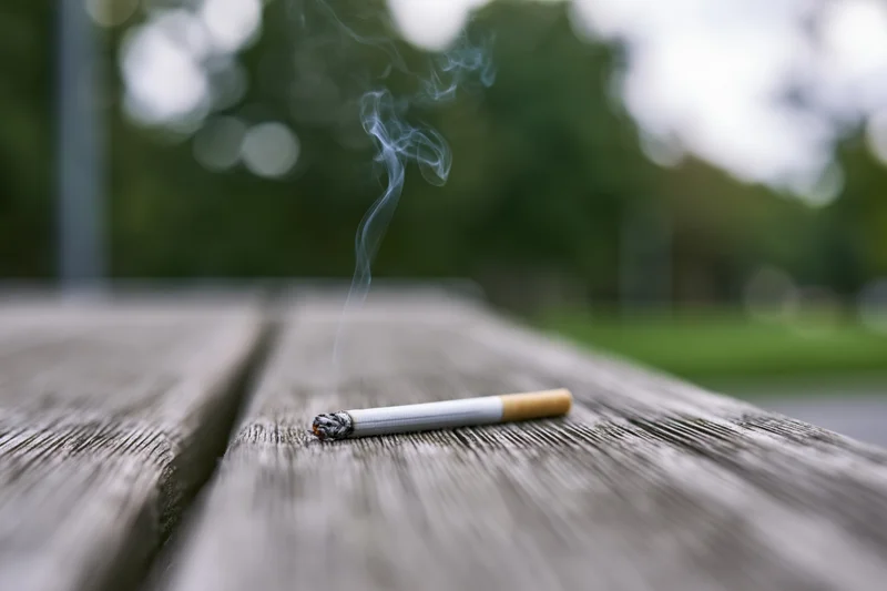 Lit cigarette with smoke resting on weathered wooden park bench outdoors