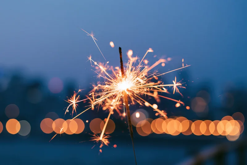 Lit sparkler glowing brightly against twilight cityscape with bokeh lights in background