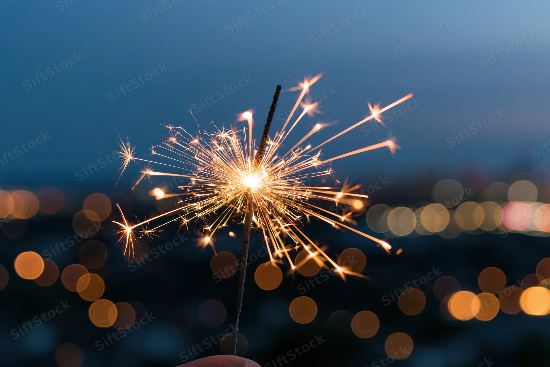Hand holding lit sparkler against twilight city skyline with bokeh lights