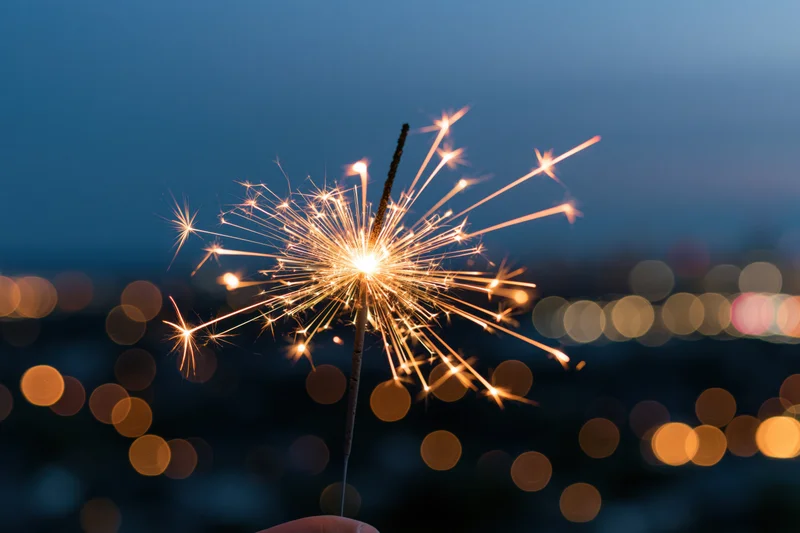 Hand holding lit sparkler against twilight city skyline with bokeh lights