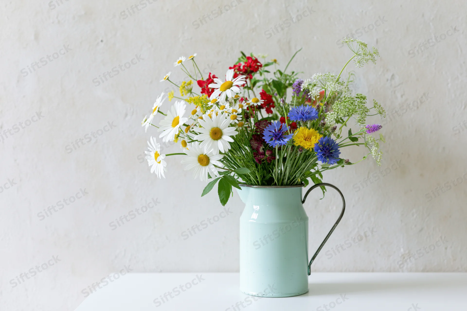 Loose bouquet of mixed wildflowers including white daisies in a mint metal pitcher on a white surface against a textured wall