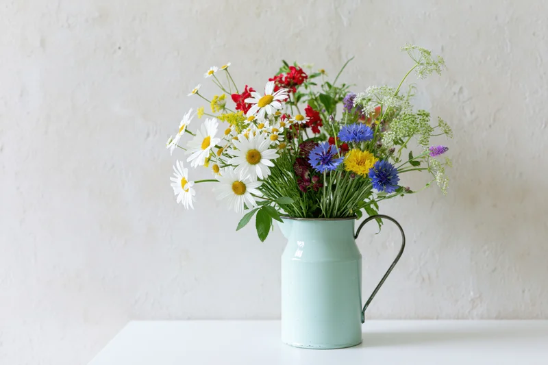 Loose bouquet of mixed wildflowers including white daisies in a mint metal pitcher on a white surface against a textured wall