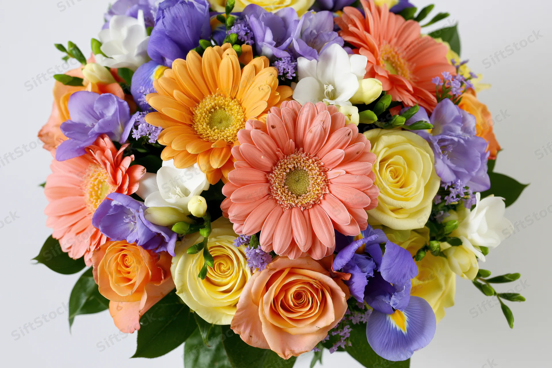 A dense mixed bouquet of gerbera daisies, roses, and irises arranged with greenery on a white background