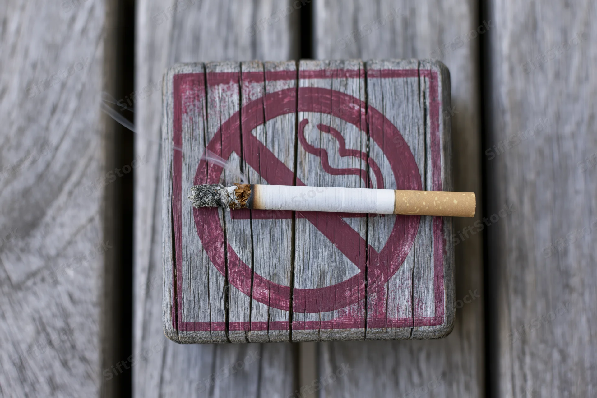 Lit cigarette resting on a no-smoking sign painted on weathered wood