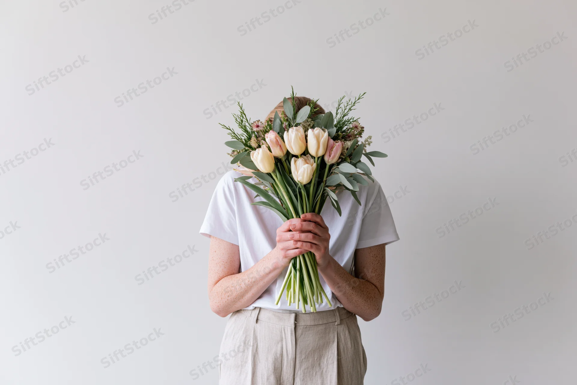 Woman holding bouquet of tulips and greenery in front of face against plain wall