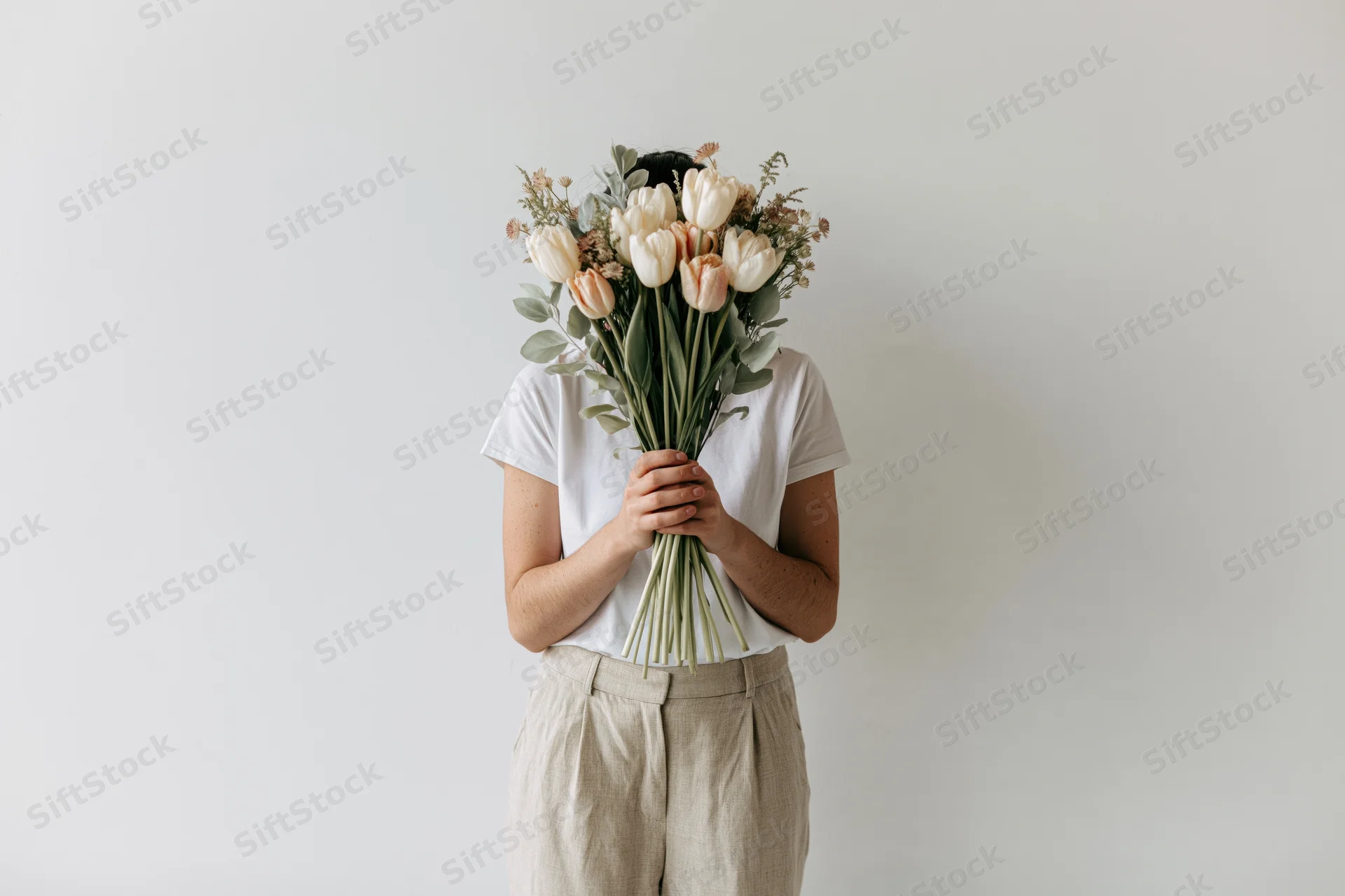 Woman holding bouquet of white tulips and eucalyptus in front of face against plain wall