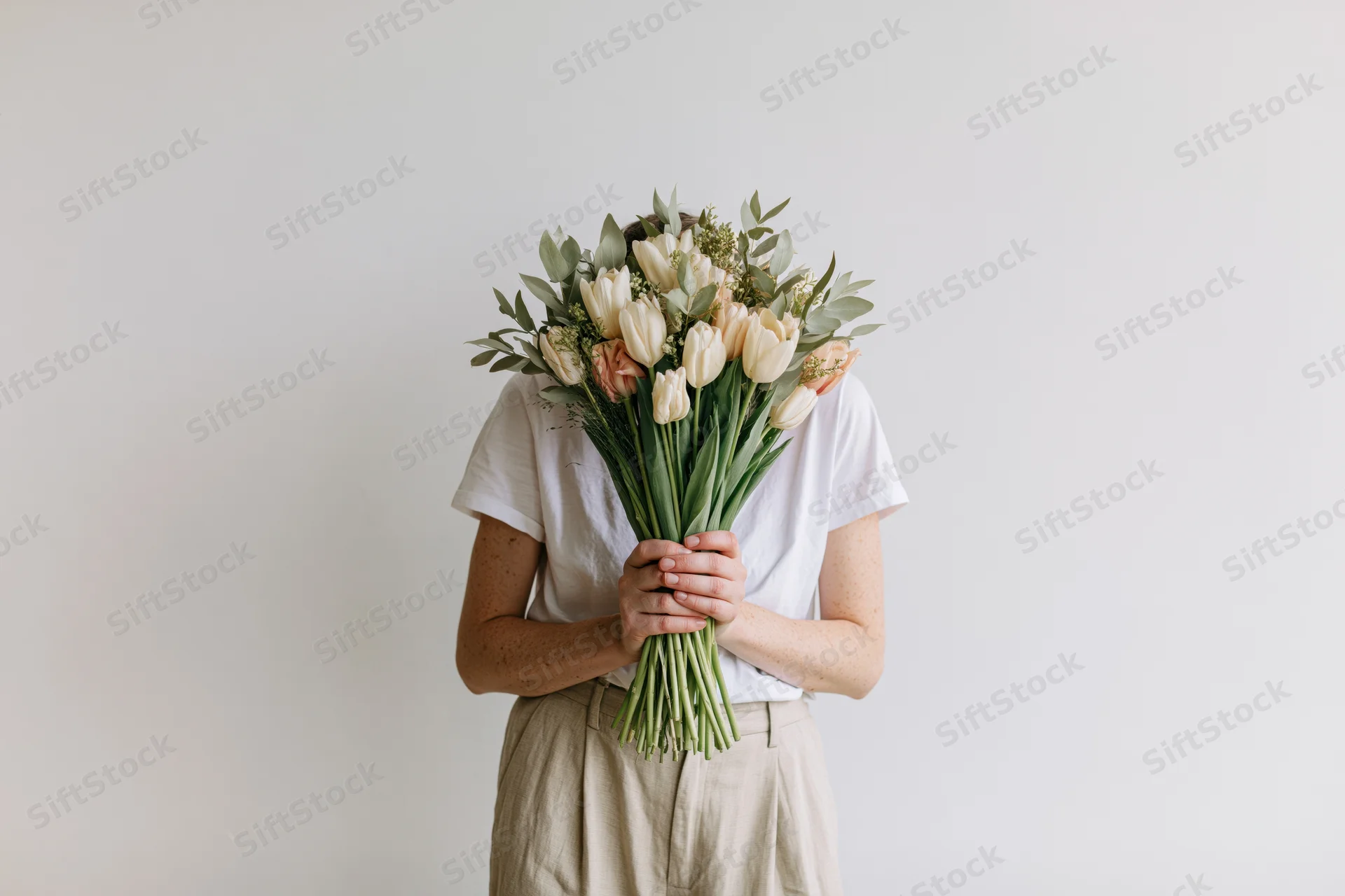Woman holding a bouquet of white tulips and greenery against a plain off-white wall