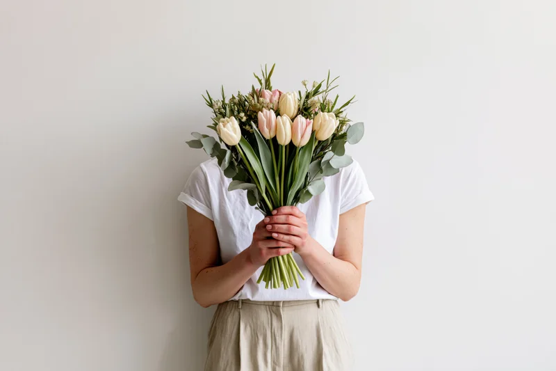 Woman holding bouquet of tulips and eucalyptus in front of face against plain off-white wall