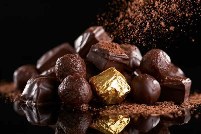 Assorted dark chocolate pralines and truffles piled on a reflective surface with cocoa powder dusting