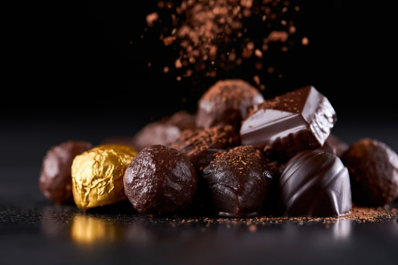 Assorted dark chocolate pralines and truffles piled on a reflective black surface with cocoa powder falling around them