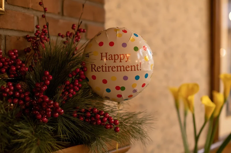 A foil balloon reading happy retirement among pine and red berries with yellow calla lilies on a brick wall background