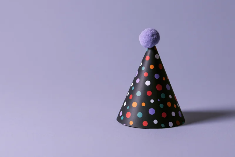 Black paper party hat with multicolored polka dots and purple pompon on plain surface