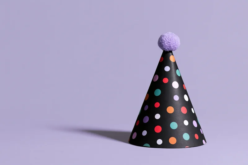 Black paper party hat with multicolored polka dots and purple pompon on lavender background