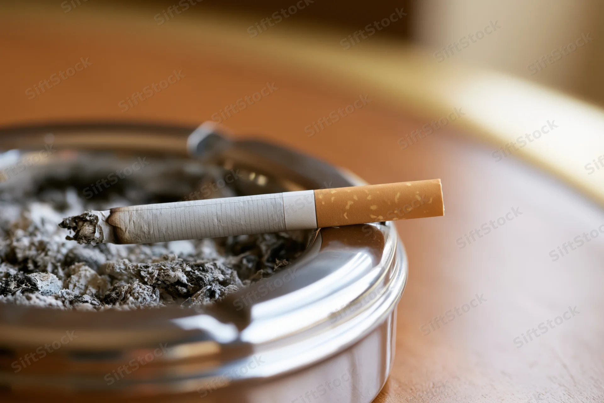 Cigarette resting on rim of chrome ashtray on wooden table
