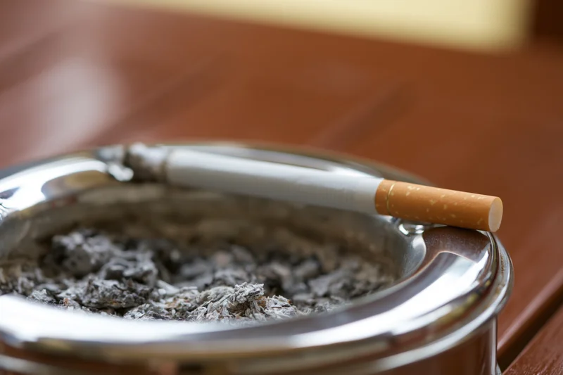 Cigarette resting on rim of chrome ashtray filled with ash on wooden table