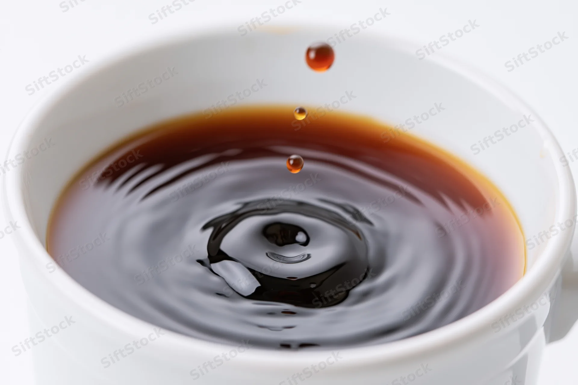 A drop of coffee suspended mid-air above a white cup with ripples forming on the surface
