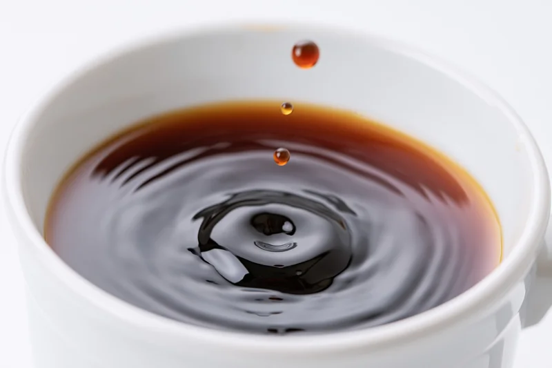 A drop of coffee suspended mid-air above a white cup with ripples forming on the surface