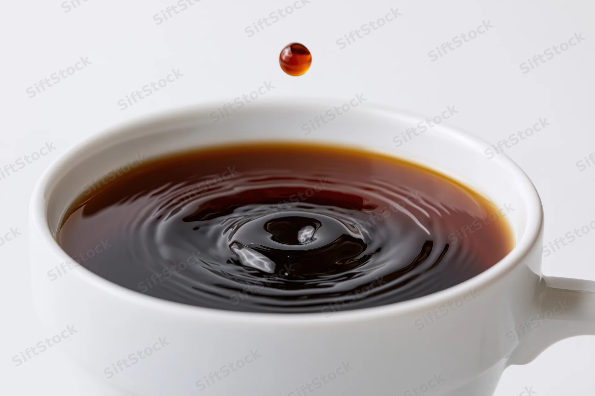A drop of coffee suspended above a white cup with ripples in the liquid