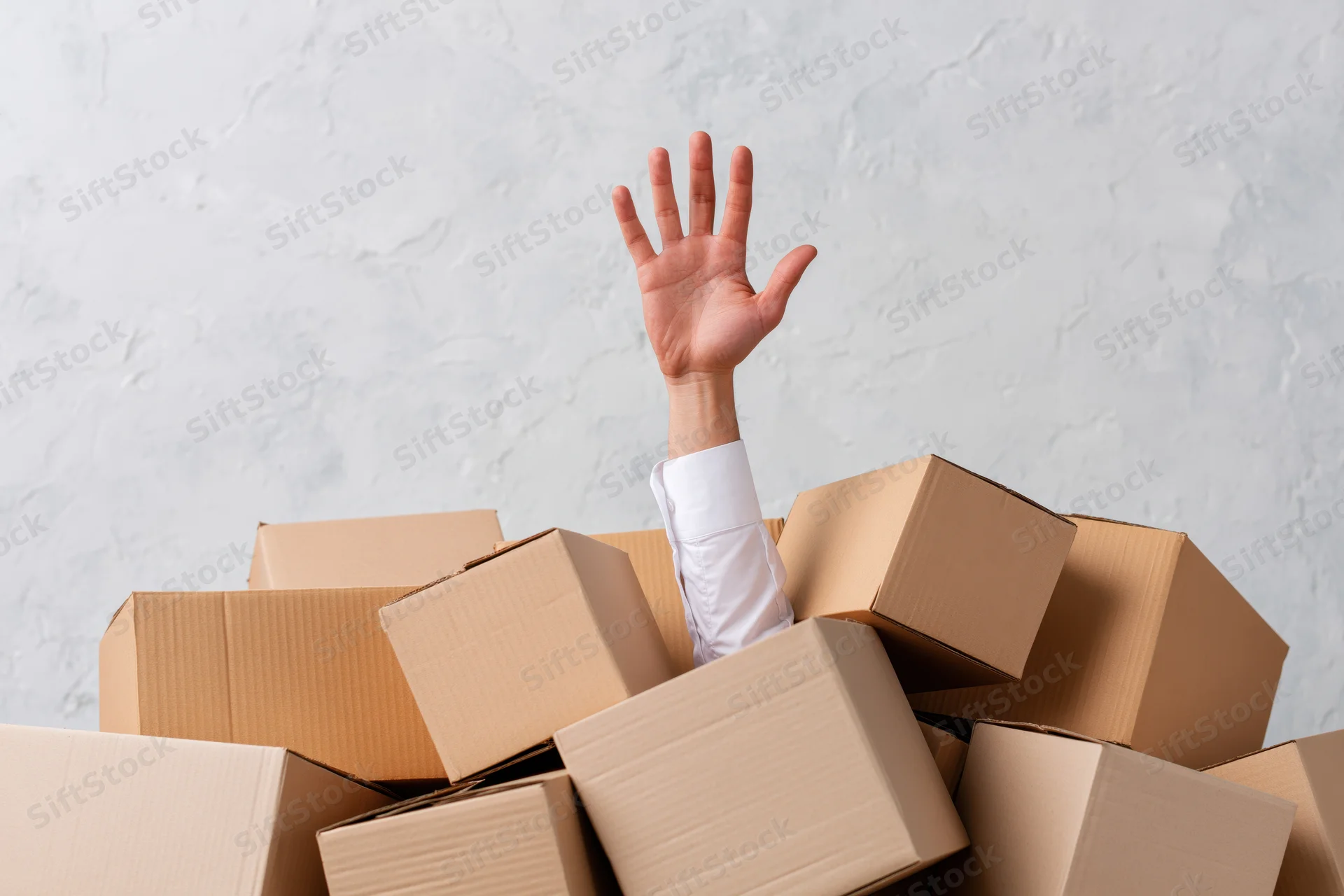 Arm reaching upward from a pile of cardboard boxes against a white wall