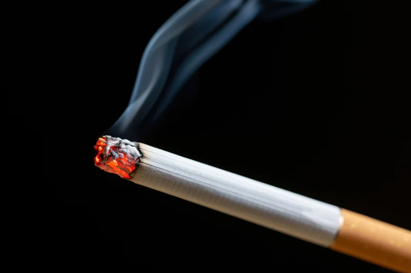 Lit cigarette burning diagonally against black background with rising smoke and glowing ember