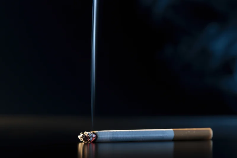 Lit cigarette with smoke rising, resting on reflective dark surface