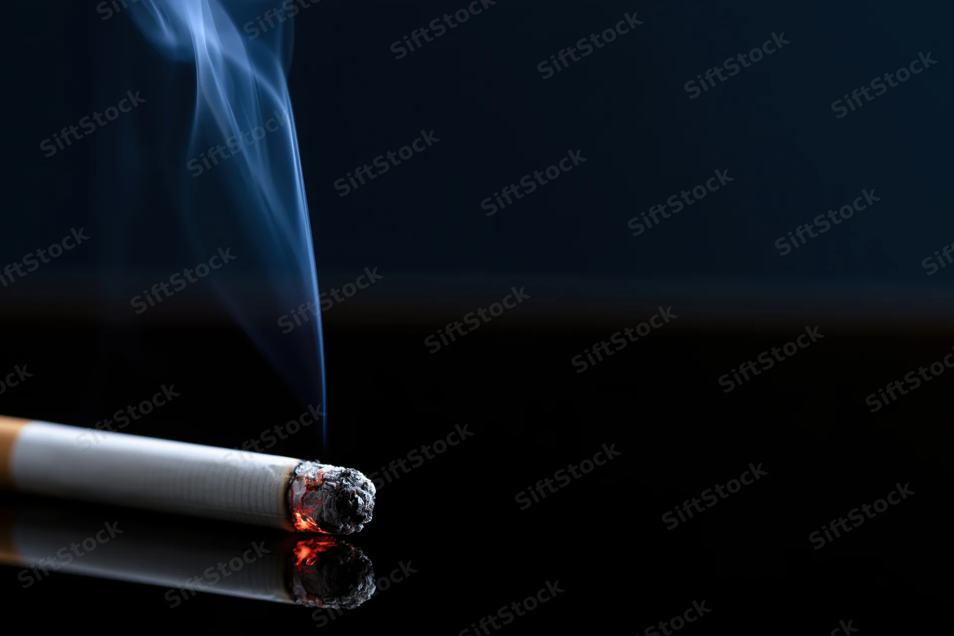 Lit cigarette burning horizontally on a reflective black surface with smoke rising and ash forming