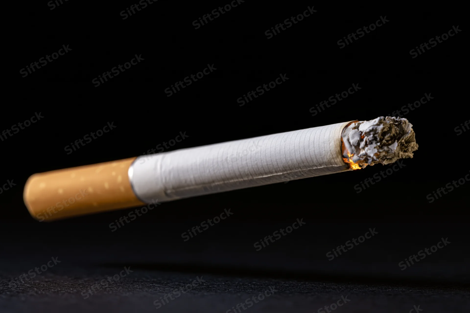 Lit cigarette burning with ash and flame suspended against black background