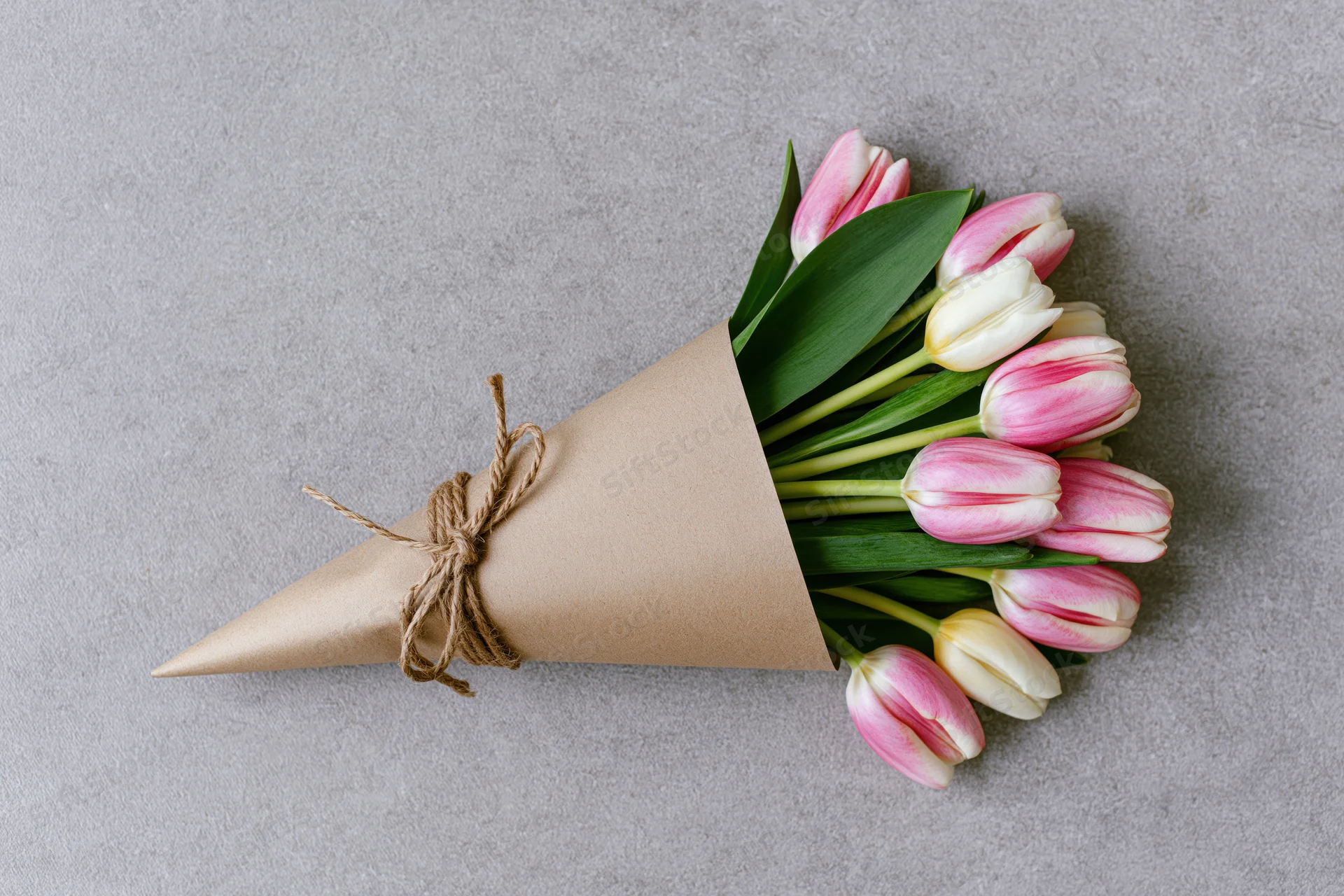 Small bouquet of pink and white tulips wrapped in kraft paper cone on gray surface