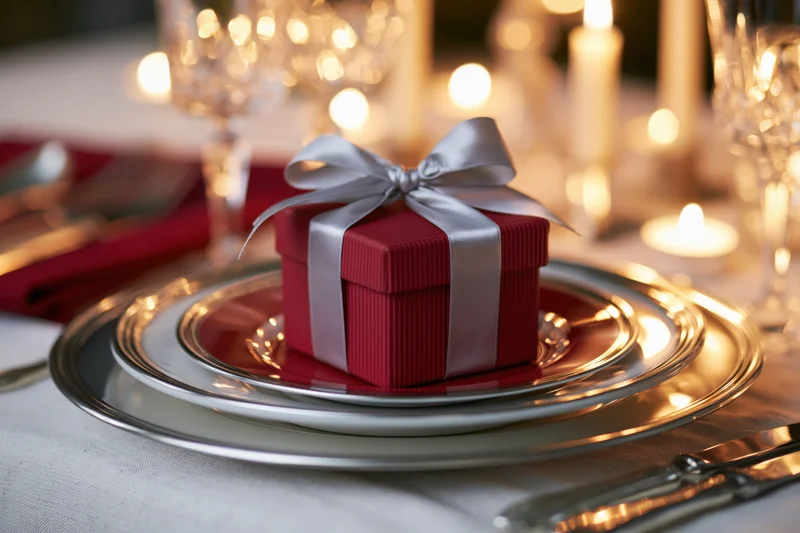 Small red ribbed gift box with silver satin bow placed on stacked plates at a formal dinner table setting with candles and glassware
