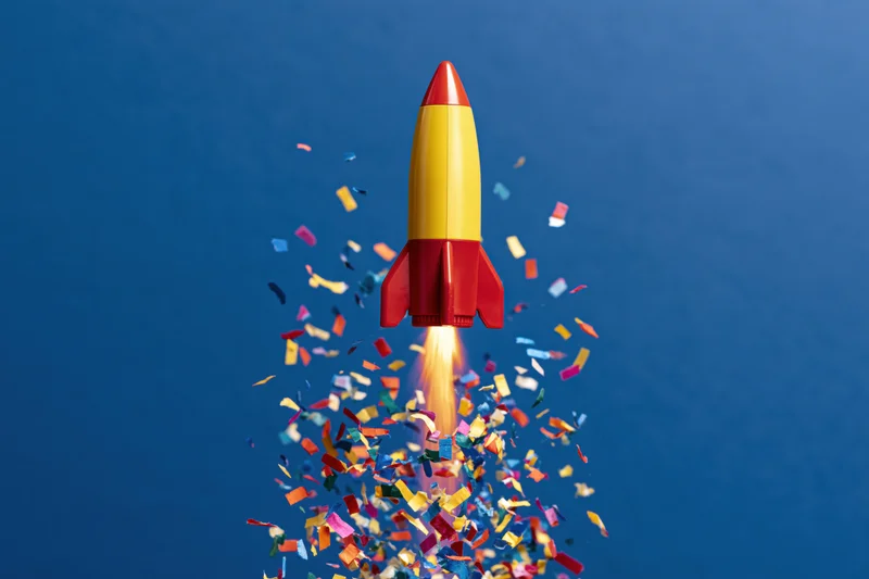 Toy rocket launching upward with confetti and flame against blue background