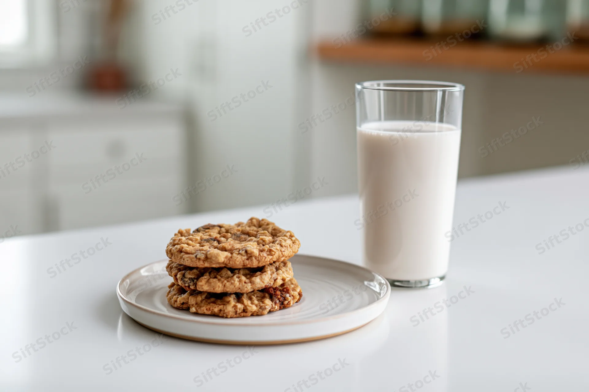Stack of oatmeal cookies on a white ceramic plate beside a glass of milk on a kitchen countertop