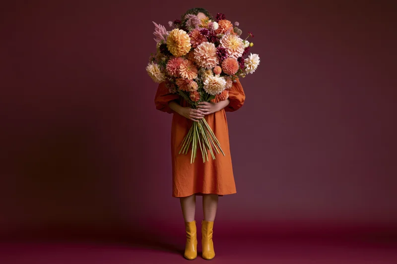 Woman holding large bouquet of autumn flowers in orange dress and yellow boots against burgundy studio background