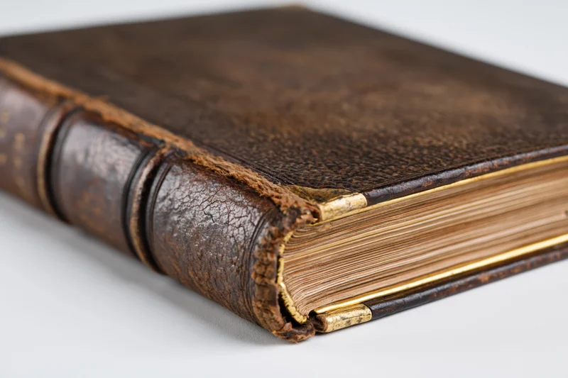 Antique leather-bound book with gilded edges resting at an angle on white surface
