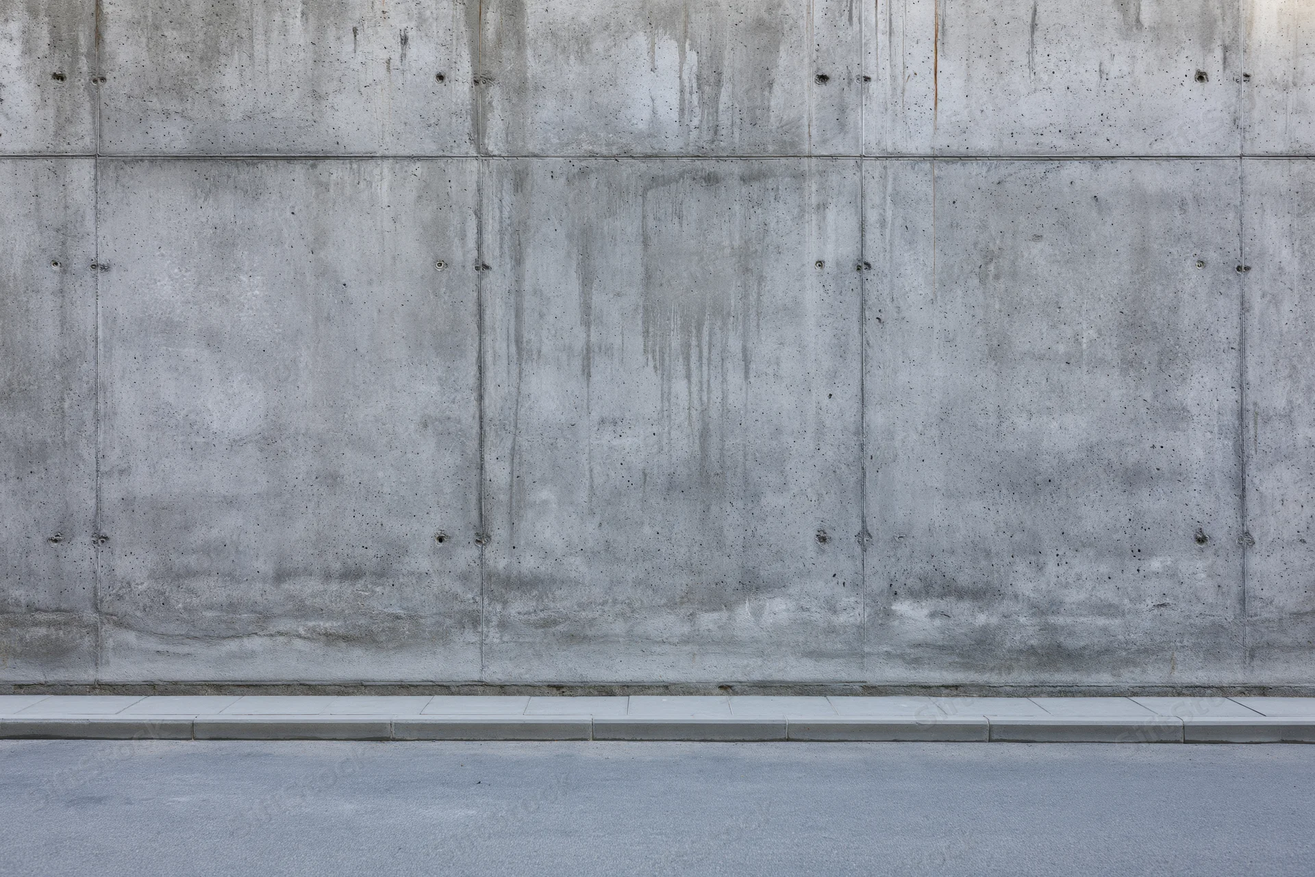 Wide view of a bare concrete wall with visible seams and stains along a sidewalk