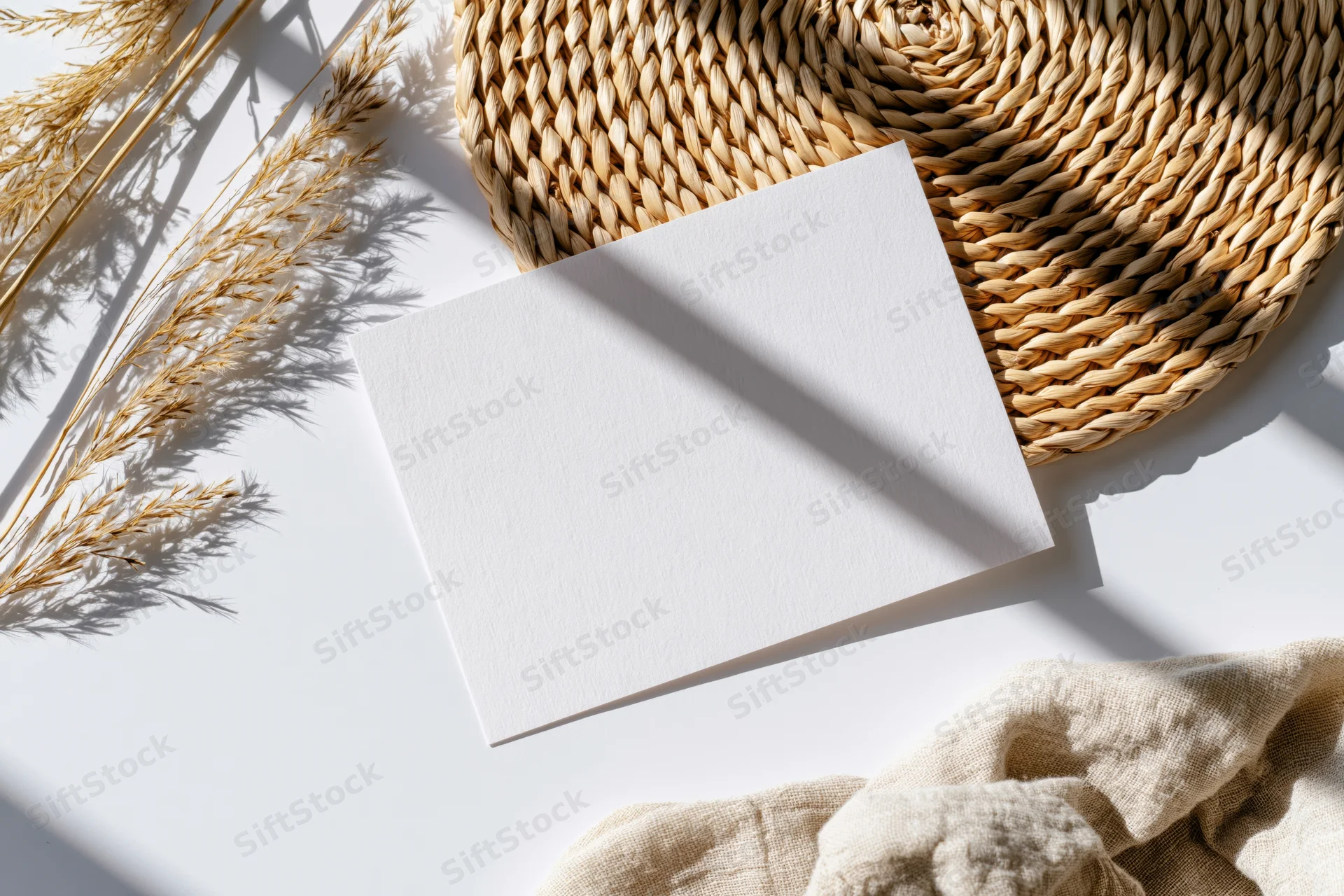 Blank white card lying flat on white surface with woven basket and dried grasses nearby