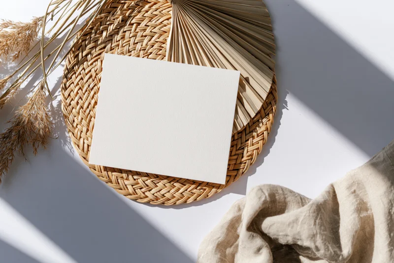 Blank white card on woven placemat with dried grass and linen fabric, flat lay on white surface