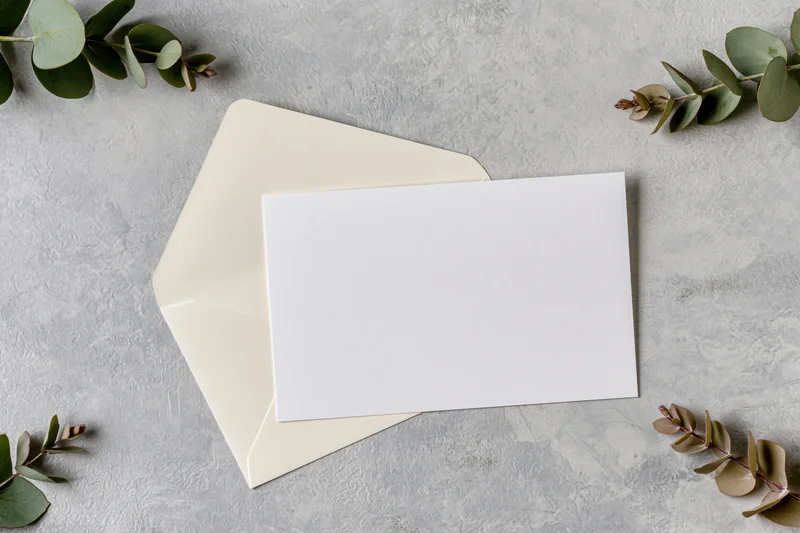 Blank white card resting on cream envelope with eucalyptus sprigs on textured gray surface