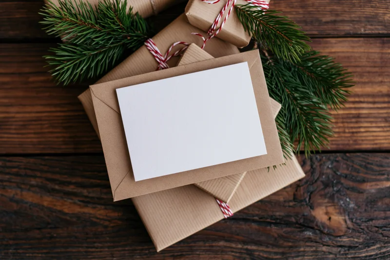 Blank white card on kraft envelope with pine branches and wrapped gifts on wooden table